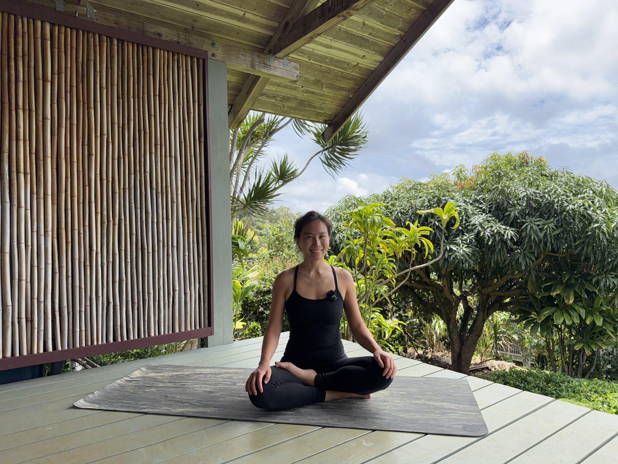 Sophia smiling, seated cross-legged on a yoga mat on an open-air deck