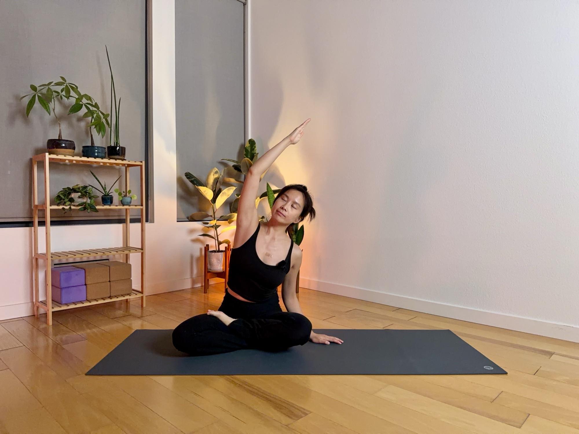 Sophia seated cross-legged on a mat in a warm, lamp-lit room, reaching one arm overhead in a side bend